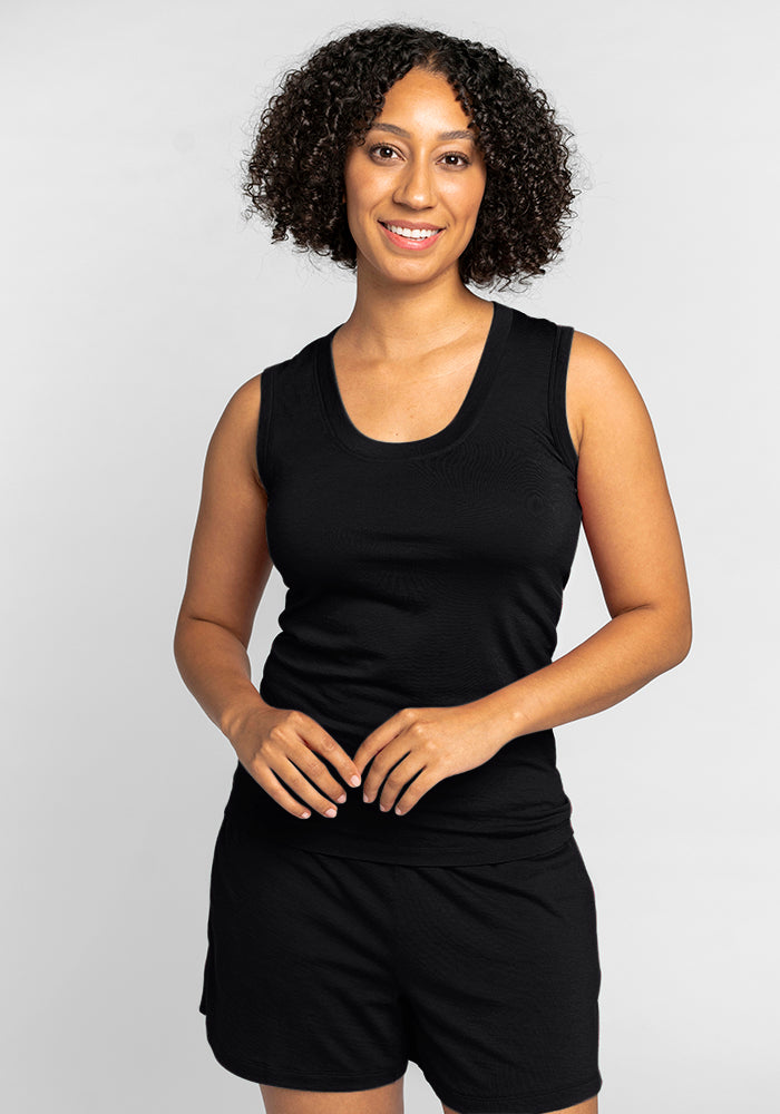 A person with curly hair smiles while standing against a plain background, wearing the Phoebe Lounge Tank in black by Woolx and matching shorts, embodying the essence of travel-friendly clothing. 