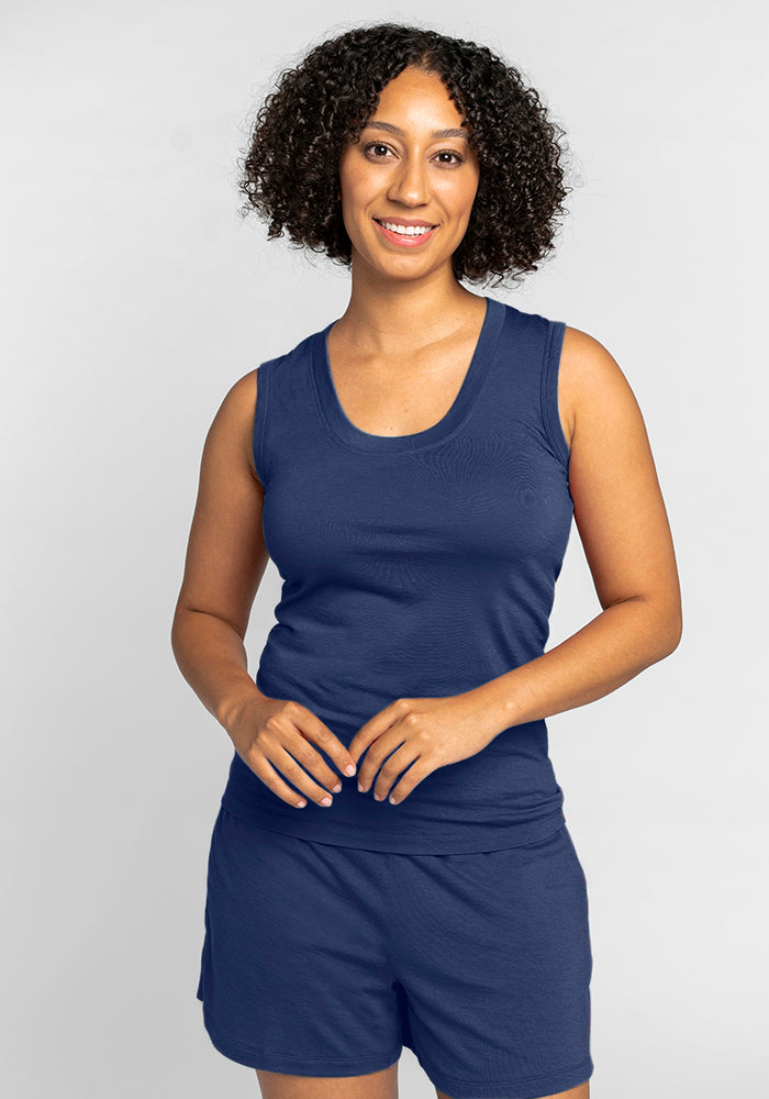 A person with curly hair is smiling, wearing a sleeveless Phoebe Lounge Tank in Starry Night and matching shorts from Woolx against a plain background. The outfit is chic and temperature-regulating, perfect for staying fresh and comfortable.