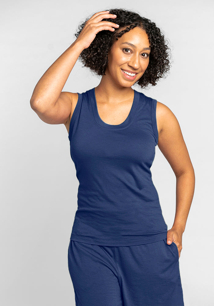 A person with curly hair is smiling while wearing the Phoebe Lounge Tank in Starry Night from Woolx and matching pants, both made from temperature-regulating Merino wool. They pose with one hand touching their hair and the other in their pocket against a plain background, exuding comfort and style. 