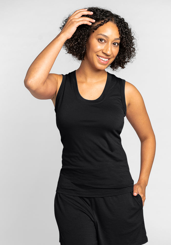 A person with curly hair smiles warmly, wearing the Woolx Phoebe Lounge Tank in black paired with black pants. Their right hand rests on their head, while the left hand is tucked in their pocket. The backdrop is plain and light-colored, ideally highlighting travel-friendly clothing.