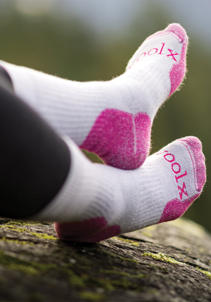 A close-up shows two feet clad in the Milo Full Cushion Cold Weather Socks in Pebble Pink, crafted from odor-resistant Merino wool, resting on a mossy rock. The brand name "Woolx" is visible on the soles. The background is slightly blurred to highlight the socks and feet.