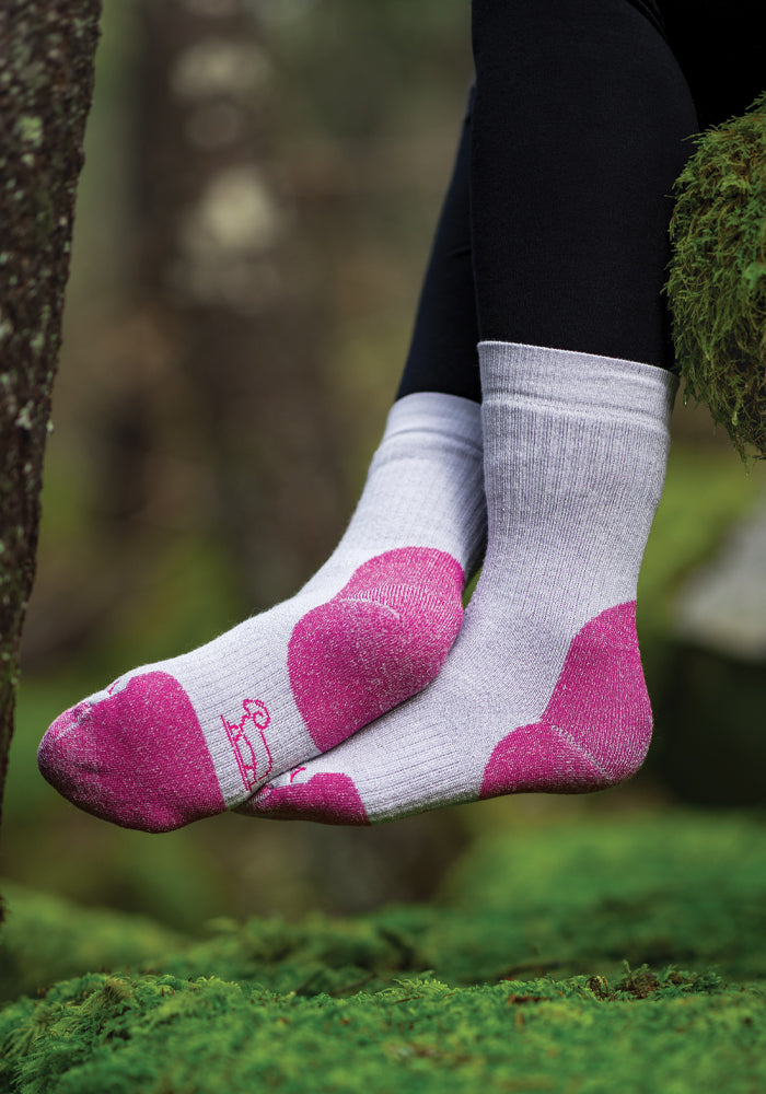 A close-up captures a person wearing Woolx's Milo Full Cushion Cold Weather Socks in Pebble Pink, crafted from moisture-wicking Merino wool. They are seated outdoors on a mossy surface, dressed in black leggings, with blurred trees visible in the background.