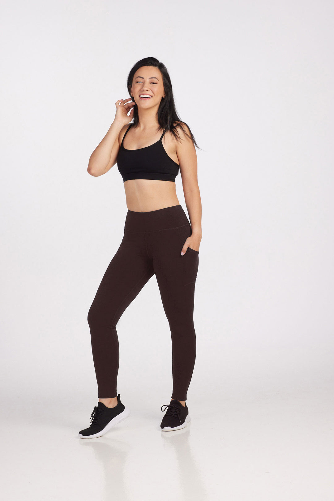 A woman dressed in a black sports bra and dark brown Woolx Piper Pocket Leggings stands against a plain white background. She is smiling and appears relaxed, with one hand touching her hair and the other in her pocket. She is wearing black sneakers, and her hair is loose.