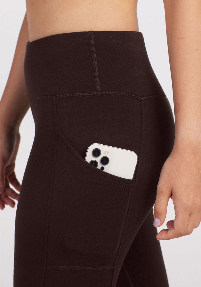 A person wearing Woolx's Piper Pocket Petite leggings in French Roast with a smartphone partially visible in the side pocket. The person has their left hand by their side and their nails are painted a light color.