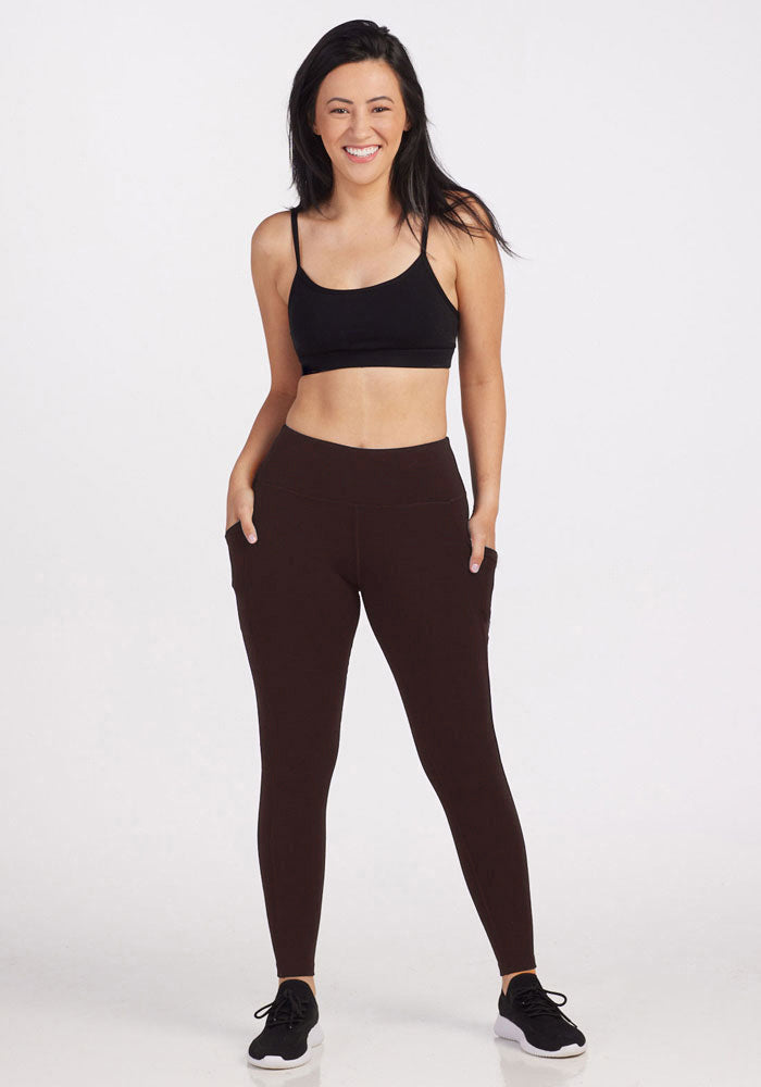 A smiling person stands against a plain background, wearing a black sports bra and dark brown high-waisted Woolx leggings specifically designed for petite body sizes, named Piper Pocket Petite in the French Roast color. Their hands are in the pockets of the leggings, and they have long dark hair, complemented by black athletic shoes.