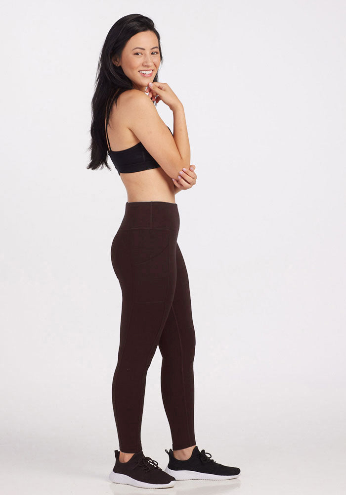 A person standing sideways to the camera, wearing a black sports bra, Woolx Piper Pocket Leggings in French Roast, and black sneakers with white soles. They have long, dark hair and are smiling while resting their chin on their left hand. The background is plain and light-colored.