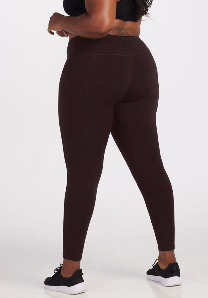 A person is wearing Woolx Piper Pocket Leggings, which are high-waisted and dark-colored with discreet pockets, paired with black sneakers that have white soles. The person is standing with their back to the camera, showcasing the fit and style of the leggings.