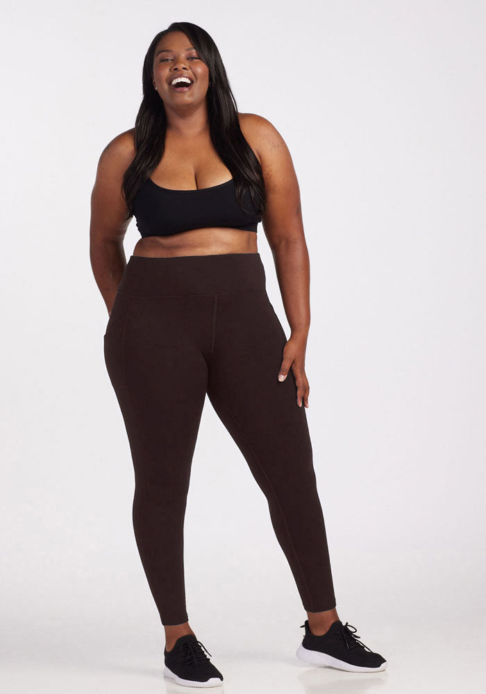 A woman posing against a plain background, smiling broadly. She is wearing a black sports bra, Woolx's Piper Pocket Leggings in French Roast, and black athletic shoes. She has long, straight hair and her hands are resting lightly on her thighs.