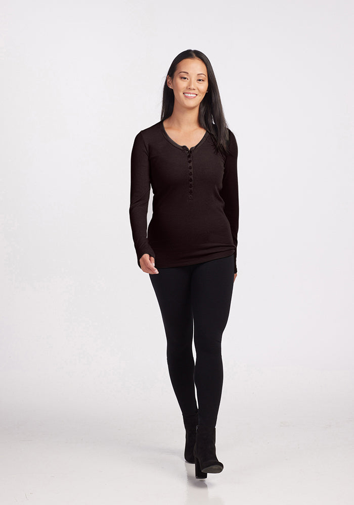 A person with long black hair is walking toward the camera, smiling. They are wearing a black Reese Ribbed Top by Woolx, black leggings, and black ankle boots. The background is plain and white.