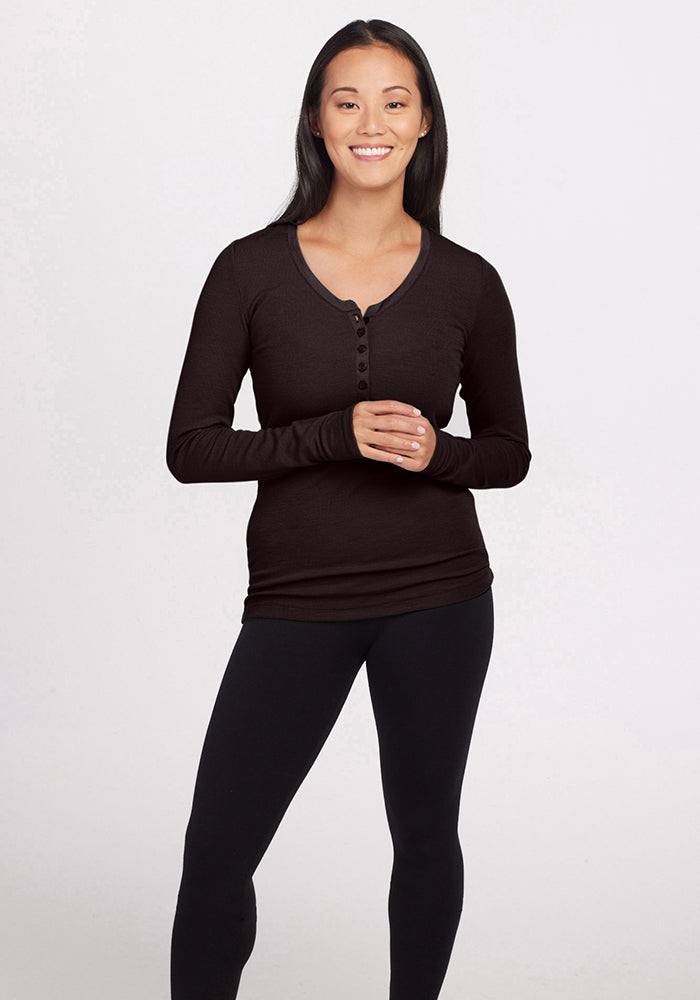 A person with long dark hair and a relaxed smile is standing against a plain background. They are wearing a Reese Ribbed Top from Woolx and black leggings. Their hands are loosely clasped in front of them. 