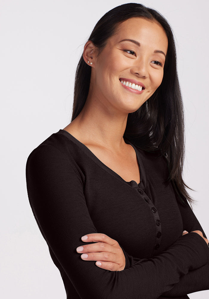 A person with long dark hair is smiling and looking off to one side. They are wearing a Reese Ribbed Top from Woolx, made from Australian Merino Wool, and have their arms crossed. The background is plain and light-colored.