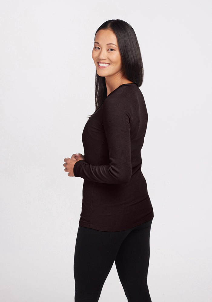 A smiling person with long black hair stands sideways, looking at the camera. They are wearing a Reese Ribbed Top by Woolx and black pants, against a plain white background.