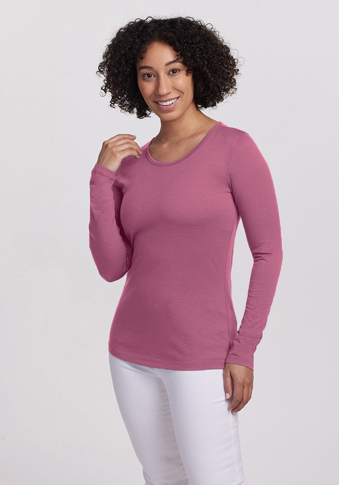 A person with curly hair is smiling, wearing a lightweight pink Remi Long Sleeve TShirt from Woolx, paired with white pants, and posing against a plain background.