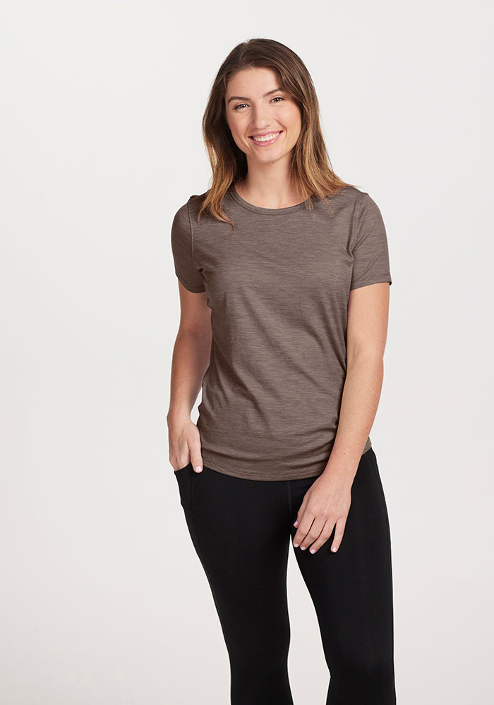 A person with long brown hair is smiling, wearing a lightweight Simply Taupe Liza Crew Neck by Woolx and black pants, standing against a plain white background.