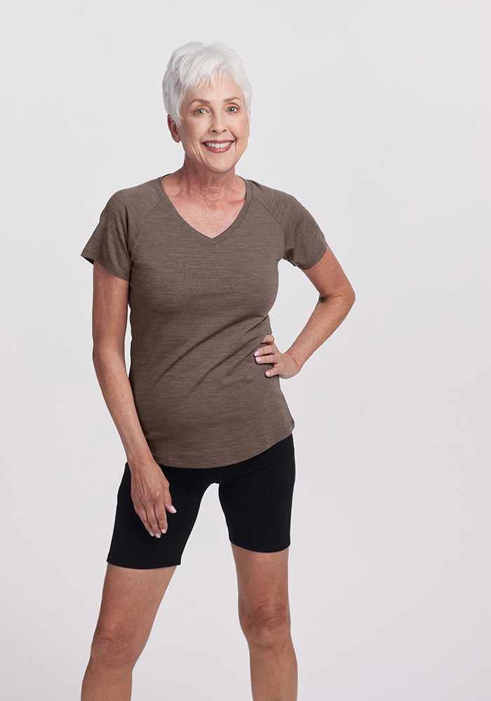 An elderly woman with short white hair confidently smiles in a Woolx Mia Short Sleeve V Neck and black shorts. She stands against a plain white background, her hand resting on her hip—a perfect look for any adventure, seamlessly combining style and comfort.