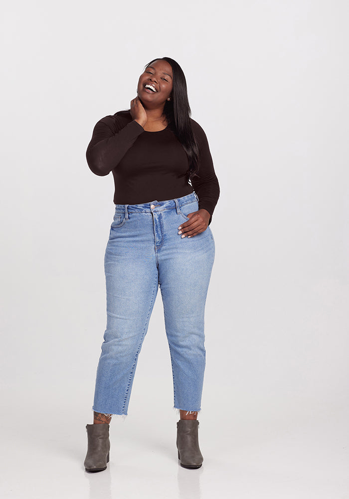 A person stands smiling and touching their neck, showcasing a chic look. They are wearing a black long-sleeve Woolx Sophia Bodysuit, light blue jeans, and gray ankle boots, against a plain white background.