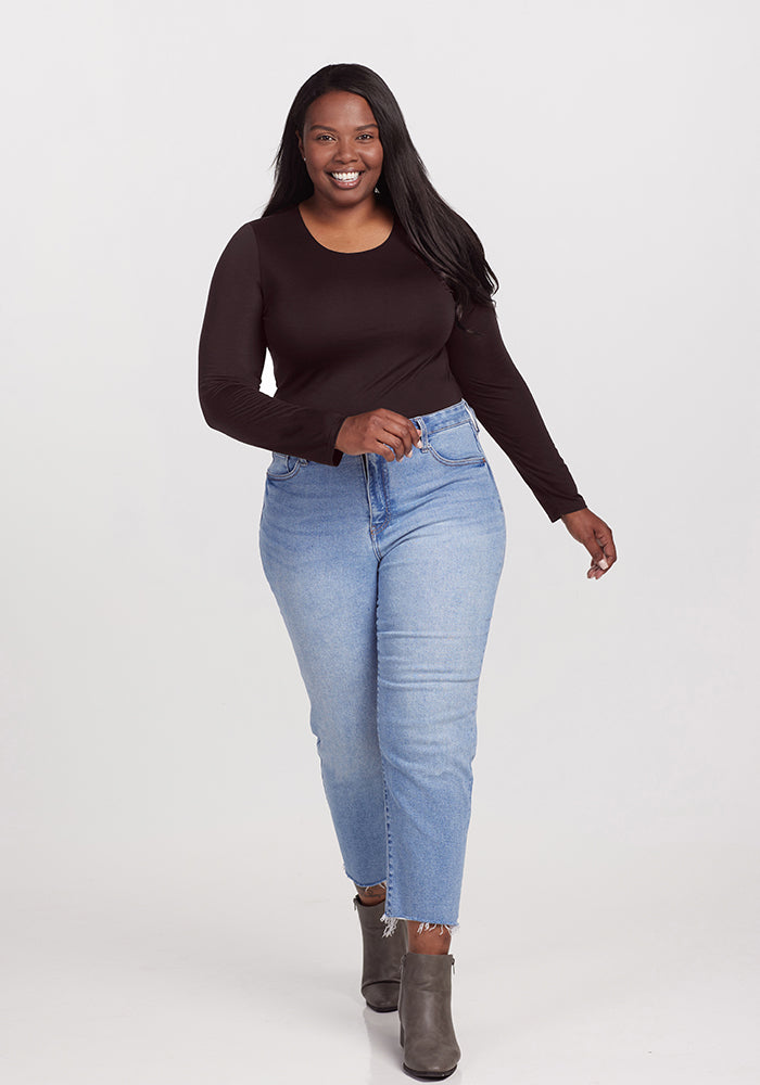 A woman with long black hair is smiling and walking towards the camera. She is wearing a black long-sleeved Sophia bodysuit from Woolx, light blue jeans, and grey ankle boots, effortlessly showcasing a chic look. The background is a plain white studio setting.