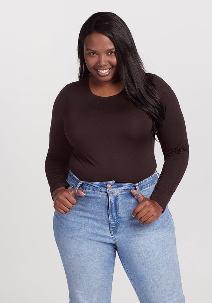 A woman with long, dark hair smiles while standing with her hands on her hips. She is wearing a chic, long-sleeve, black Woolx Sophia Bodysuit and high-waisted, light blue jeans. The background is plain white. 