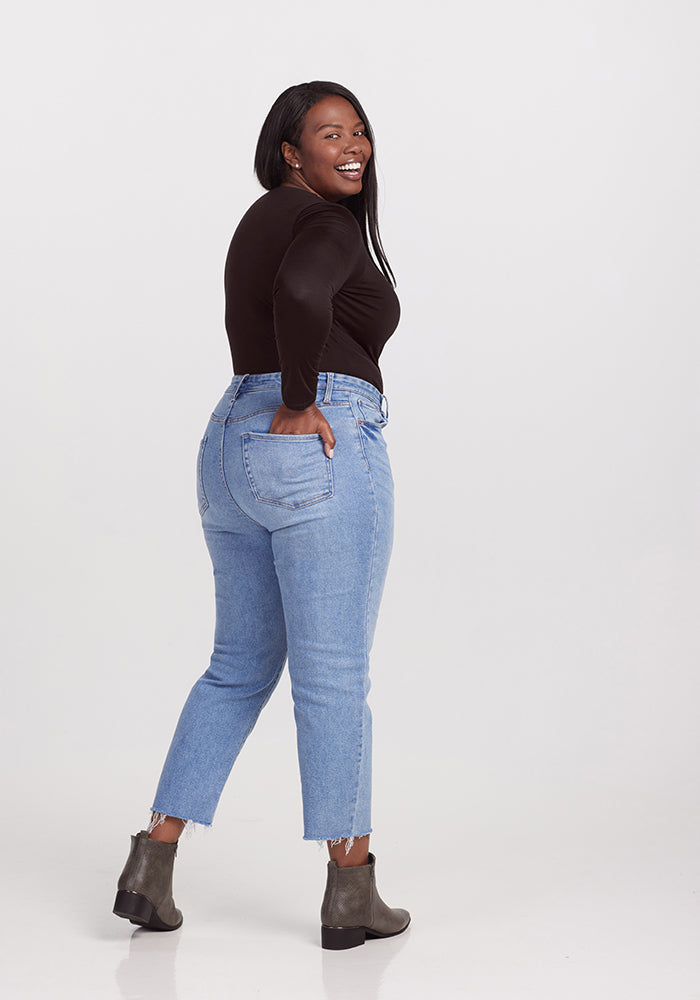 A woman with long dark hair, wearing a chic black Woolx Sophia Bodysuit, light blue jeans, and gray ankle boots, stands sideways with one hand on her hip, smiling confidently against a plain white background.