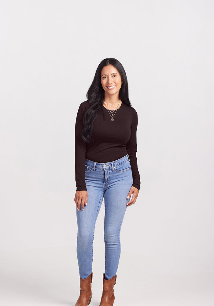 A woman with long, dark hair stands against a plain background, smiling at the camera. She is wearing a black Woolx Sophia Bodysuit, light blue jeans, and brown ankle boots. She has one arm relaxed at her side and the other bent slightly at the elbow. Her chic look is effortlessly stylish.