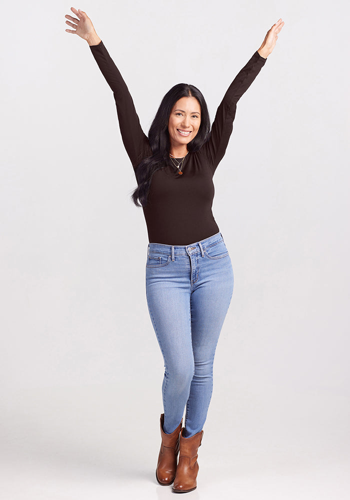 A woman with long black hair stands with her arms raised in the air, smiling. She is wearing a chic Woolx Sophia Bodysuit, light blue jeans, and brown ankle boots. The background is plain and light-colored.