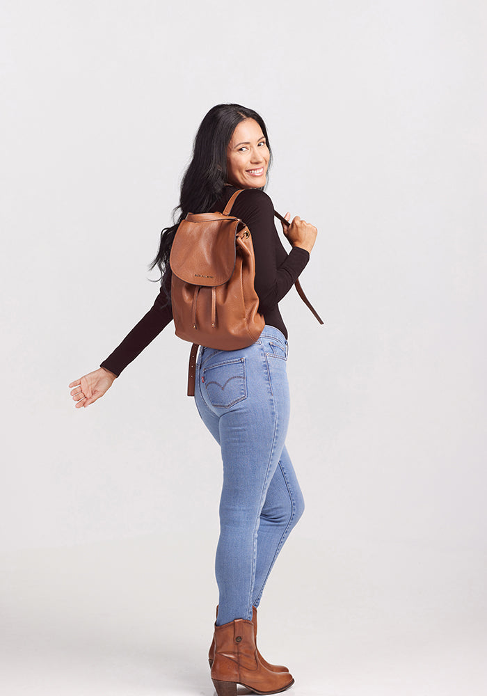 A woman with long dark hair smiles while looking over her shoulder. She is wearing a chic black Woolx Sophia Bodysuit, light blue jeans, brown ankle boots, and a brown leather backpack. She stands against a plain white background.