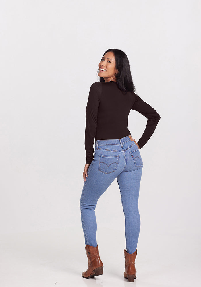 A woman with long, dark hair is smiling and looking over her shoulder at the camera. She is wearing the chic Sophia Bodysuit by Woolx, blue jeans, and brown ankle boots. The background is plain and white.