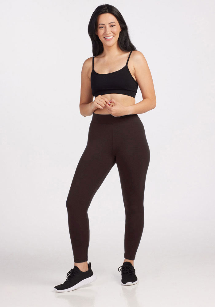 A woman with long dark hair stands smiling against a plain white background. She is wearing a black sports bra, the Stella Petite leggings in French Roast by Woolx, and black sneakers. She has a relaxed stance with her arms casually held in front of her, showcasing the comfortable fit of her outfit.