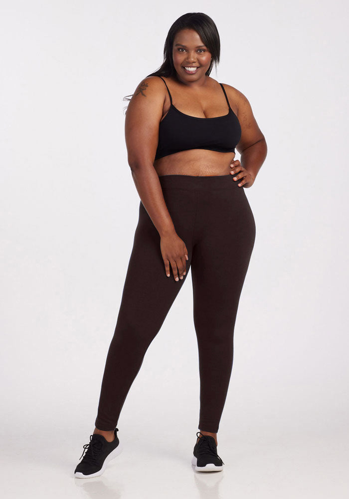 A smiling woman stands confidently with one hand on her hip. She is wearing a black sports bra, French Roast Stella Leggings from Woolx, and black athletic shoes. She has long dark hair and visible tattoos on her upper arm. She poses against a plain white background.