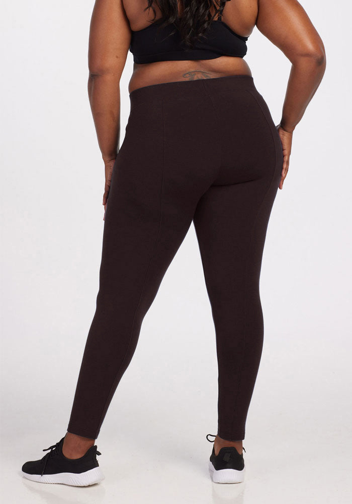 A person is seen from the back showcasing Woolx athletic wear, including a black sports bra and Stella Leggings in French Roast. They are standing against a plain white background, with their hands resting on their thighs while also wearing black sneakers.