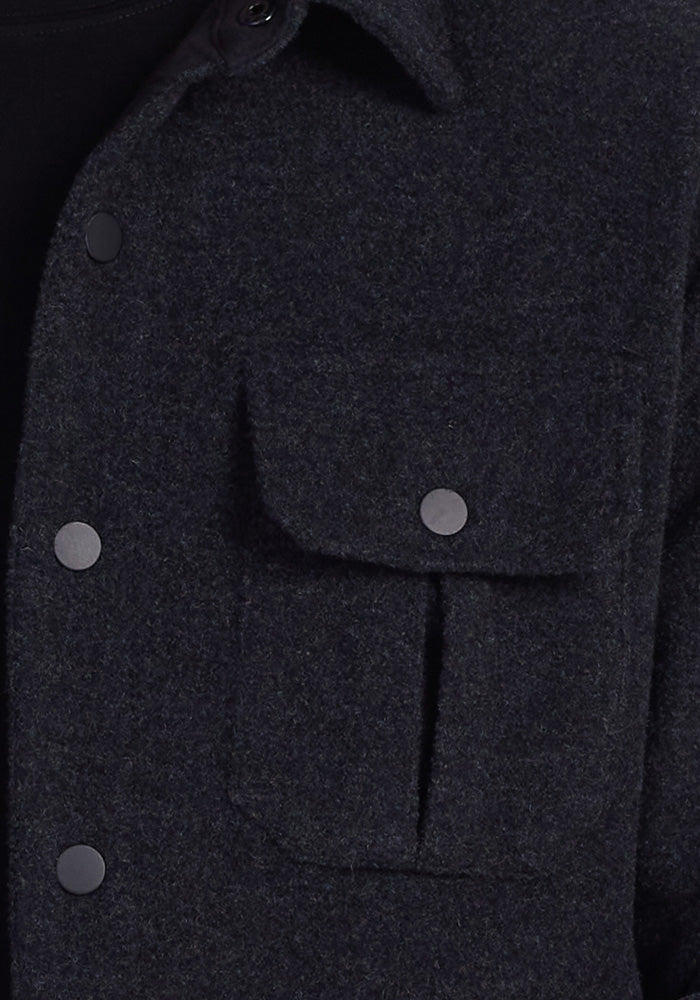 Close-up of the Wilder Shirt Jac in Carbon Black by Woolx. This ultra-functional dark gray woolen jacket features a pointed collar and a chest pocket, with metallic button closures on both the pocket and along the front. The texture of the durable knit fabric is clearly visible, highlighting its softness and warmth.