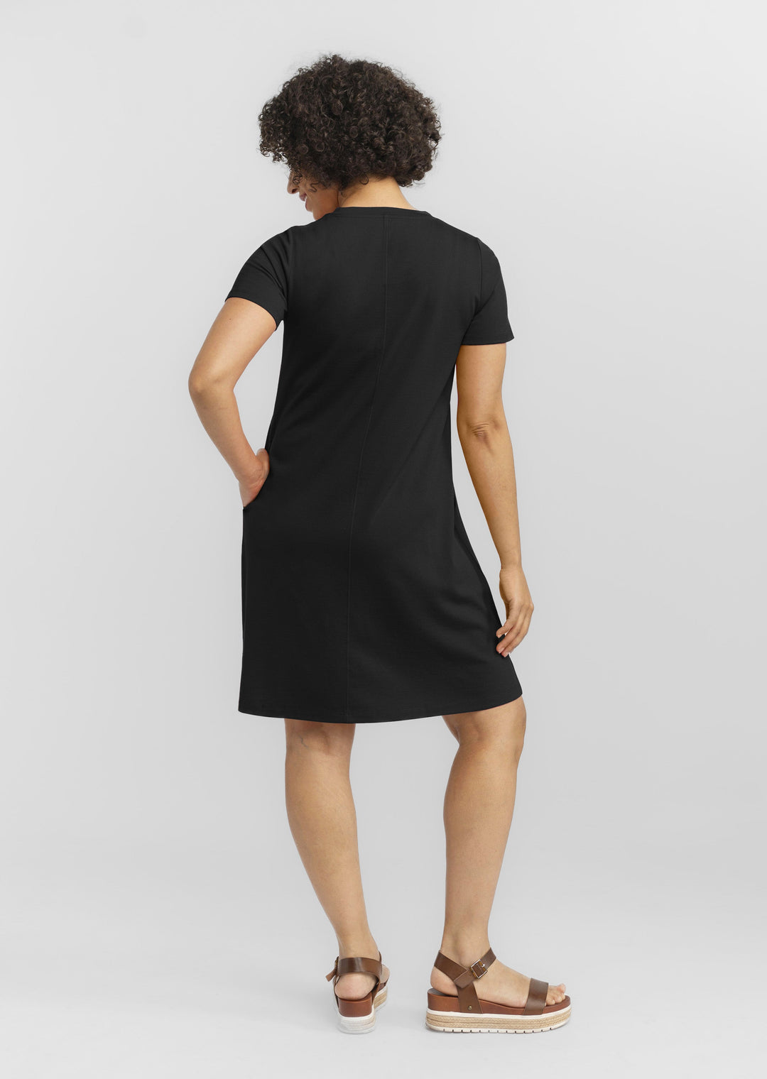 A person with curly hair is facing away, wearing Woolx's short-sleeved, knee-length Georgie Dress in black, made of merino wool. They are standing with one hand on their hip and the other relaxed by their side, complemented by brown platform sandals. The background is plain white.