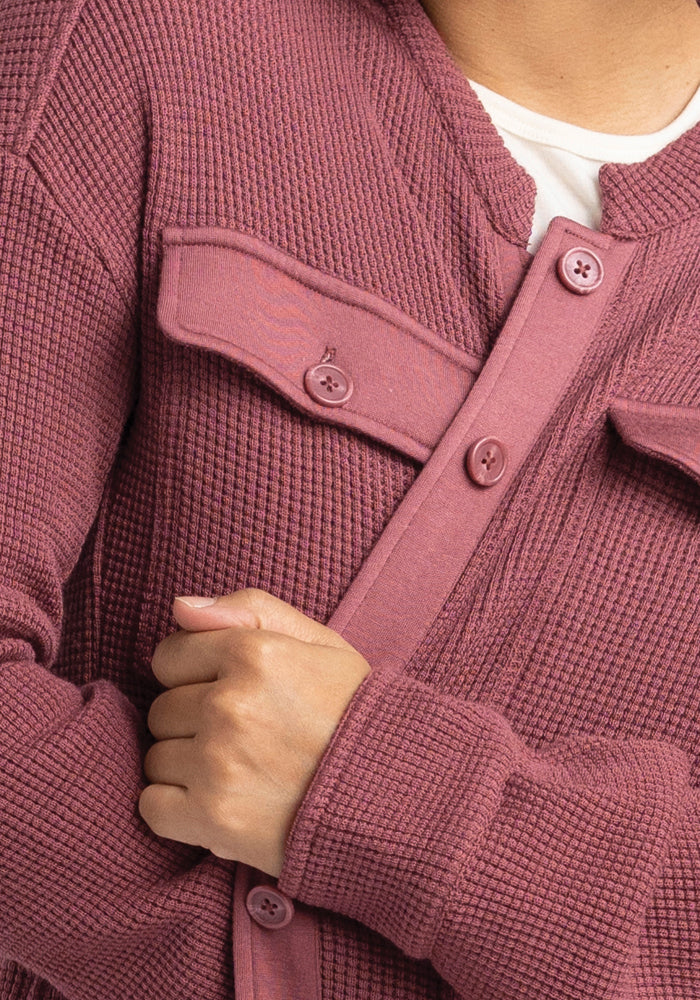 A close-up of a person wearing the Oaklynn Waffle Shacket in a rich Wild Ginger hue from Woolx. This textured layer, crafted from luxurious Merino Wool, features buttoned pockets. The individual’s arms are crossed over the shacket, which is stylishly layered over a crisp white shirt.