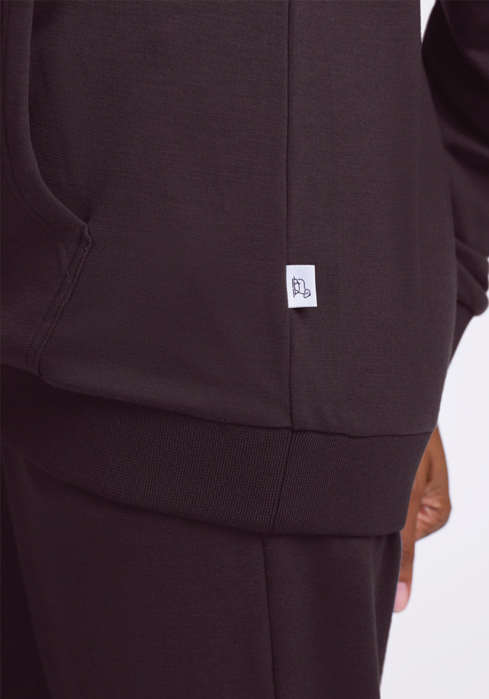 A person wears a Woolx Avery Hoodie in Chocolate Plum from the WoolAire Collection. The image highlights the side of this Merino wool creation, showcasing a small tag with a subtle logo and a partially visible hand near the bottom.