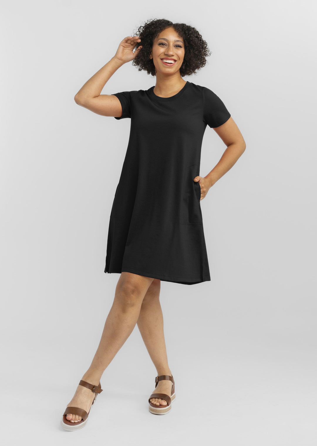 A woman with curly hair is smiling and posing confidently against a plain backdrop. She is wearing a short-sleeved, black Georgie Dress from Woolx and brown sandals with a slight heel. One hand is raised near her head while the other rests in a pocket, exuding cheerfulness.