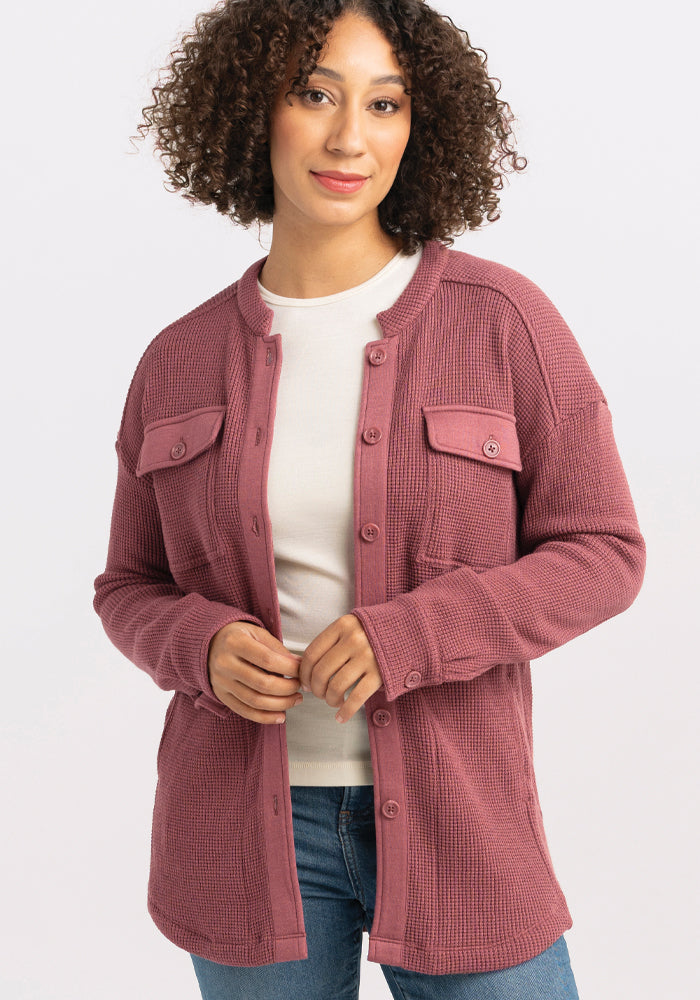 A person with curly hair wearing a Woolx Oaklynn Waffle Shacket in Wild Ginger over a white shirt stands against a plain background. They are smiling and have their hands gently clasped in front.