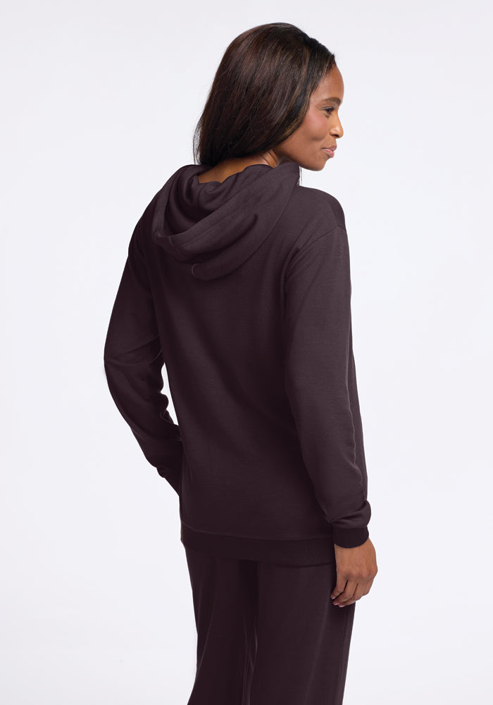 A person wearing a chocolate plum Avery Hoodie by Woolx, from the WoolAire Collection, made with soft Merino wool, pairs it with pants while facing slightly away and looking to the side. The hood is down, giving the outfit a casual and comfortable vibe against a plain white background.