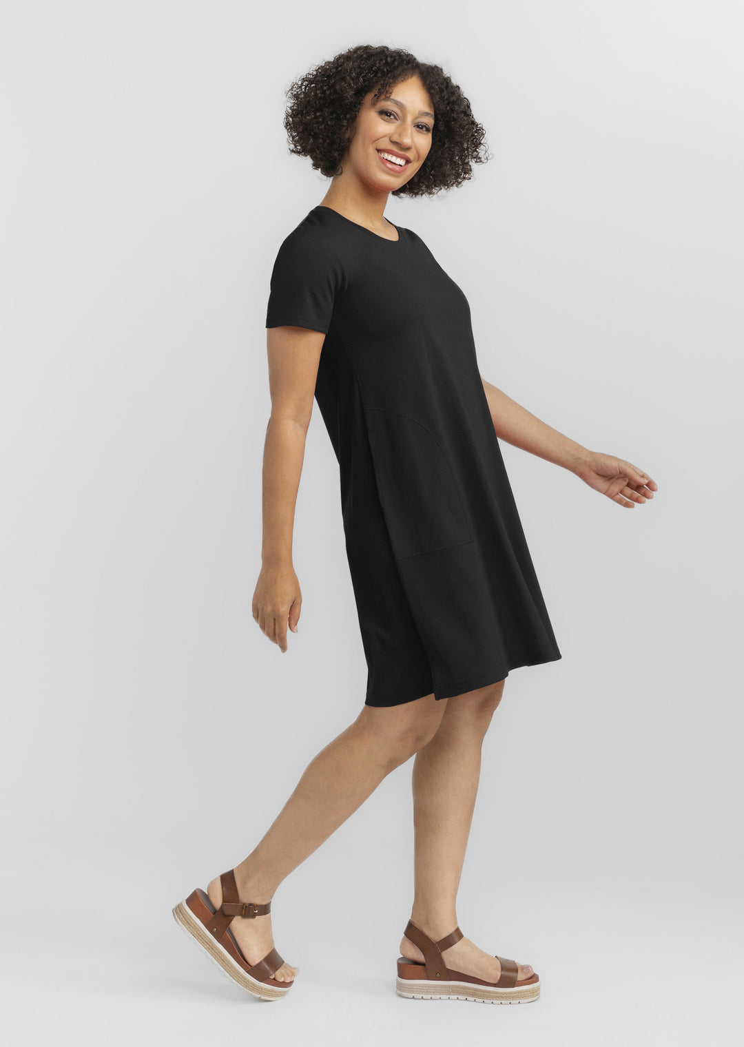 A person with curly hair is smiling and walking while wearing a Woolx Georgie Dress - Black, featuring a relaxed fit, paired with brown strappy sandals. The background is plain and light-colored.