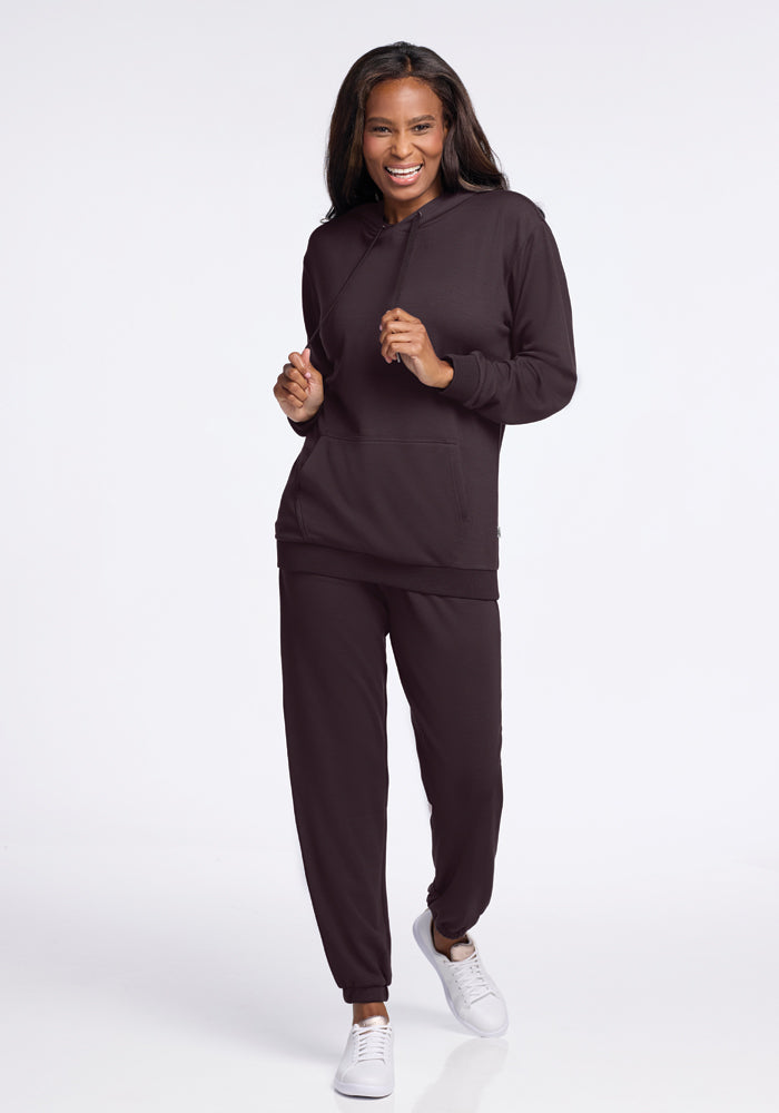 A person is smiling and posing in a chocolate plum Woolx Avery hoodie tracksuit with jogger pants. They are wearing white sneakers and standing against a plain white background, embodying the comfort of the WoolAire Collection.