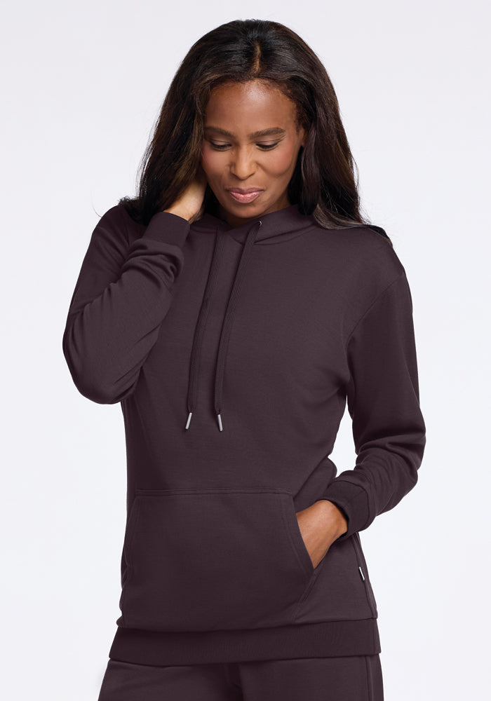 A person wearing an Avery Hoodie in Chocolate Plum from Woolx stands against a plain background. Their long dark hair frames their face as their right hand rests on the back of their neck, while the left is tucked into the cozy Merino wool pocket, looking down thoughtfully.