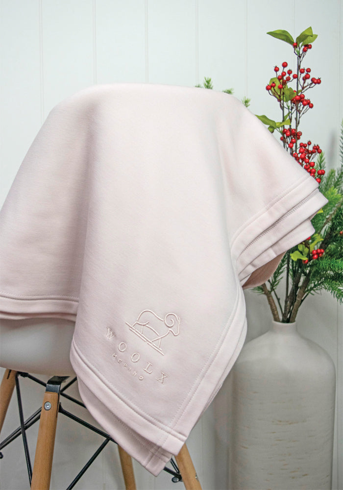 An Almost Mauve Placid Merino Blanket by Woolx, featuring an embroidered logo of a sheep and the word "WOOLY," is elegantly draped over a chair, radiating cozy vibes. In the background, a vase filled with greenery and red berries enhances the tranquil setting against a white paneled wall.