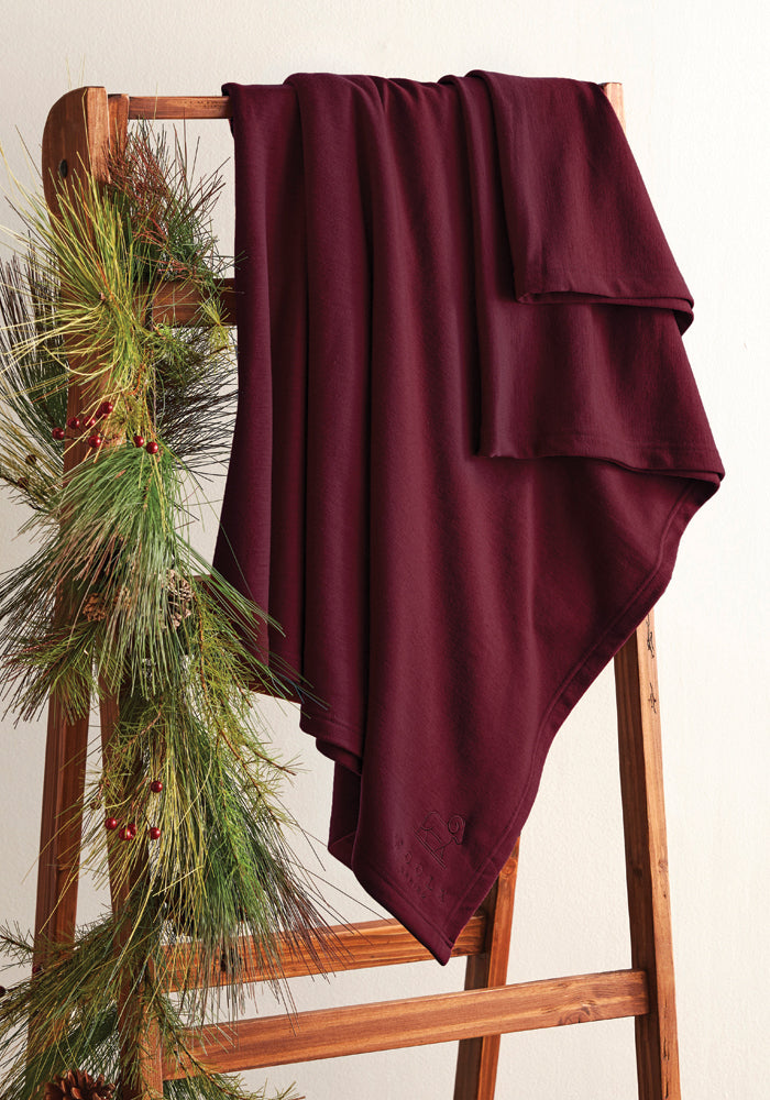 A Placid Merino Blanket in Windsor Wine, crafted by Woolx, is elegantly draped over a wooden ladder adorned with pine branches and red berries, creating a cozy and festive display against a light-colored wall.