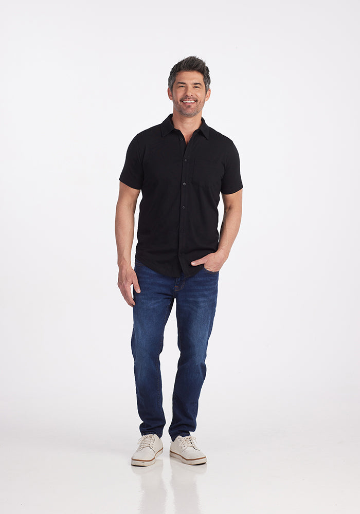 A man stands smiling against a plain white backdrop, wearing a stylish black Endi Full Button Down Shirt by Woolx paired with blue jeans and white sneakers. His arms are relaxed by his sides, radiating casual elegance.