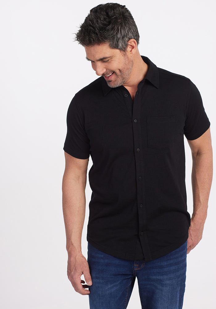 A person wearing a Woolx Endi Full Button Down Shirt in black and blue jeans looks down and smiles against a plain white background.