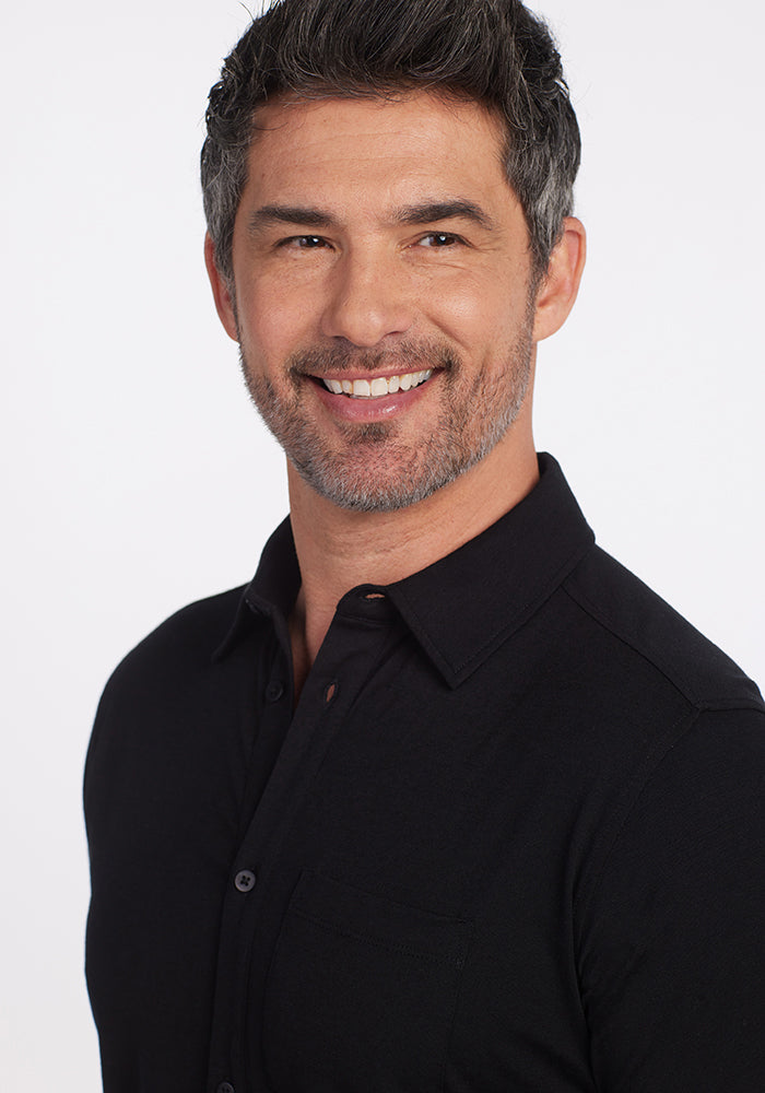 A smiling man with short, grayish hair and a beard wears a Black Endi Full Button Down Shirt from Woolx. He stands against a plain white background.