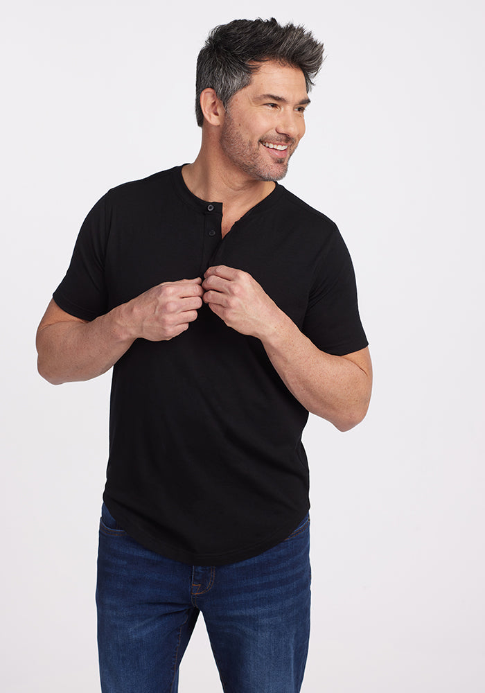 A man wearing the Bronx Henley in black from Woolx pairs it with blue jeans, smiling as he buttons up. His short dark hair with gray streaks and beard create an effortlessly chic appearance. The plain white background accentuates his style, evoking the laid-back yet sharp vibe of the Bronx. 