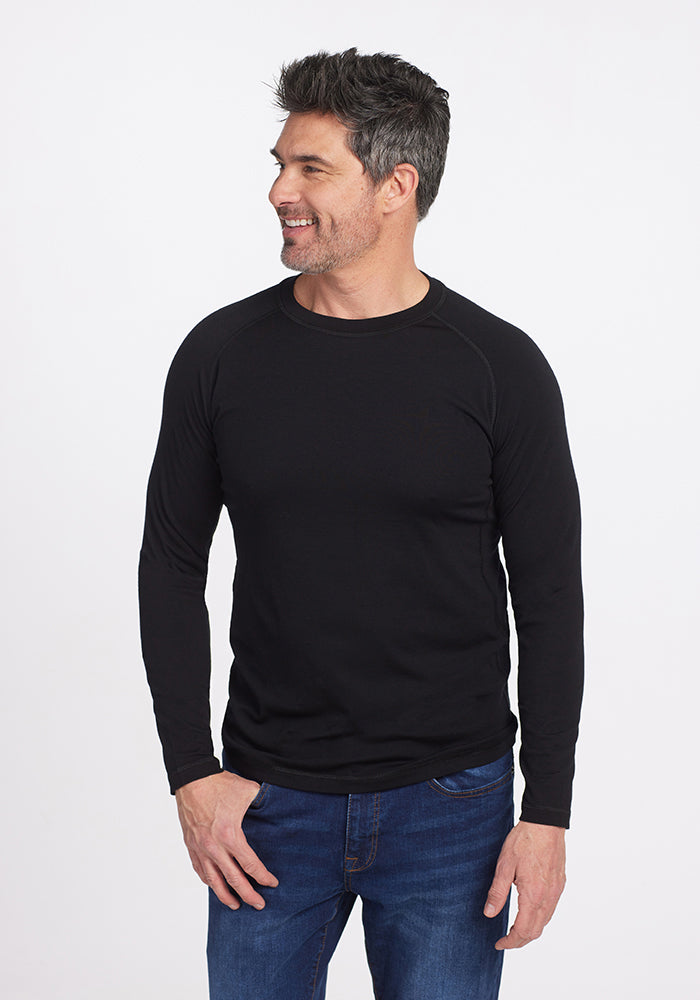 A man with short dark hair smiles and looks to the side, wearing a Woolx Essential Tee - Black and blue jeans against a plain white background.