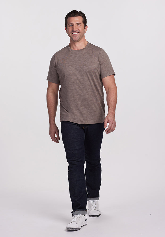 A person is walking confidently, wearing an Endurance Tee in Simply Taupe from Woolx, paired with dark jeans and white sneakers. The performance gear stands out against the plain, light-colored background.