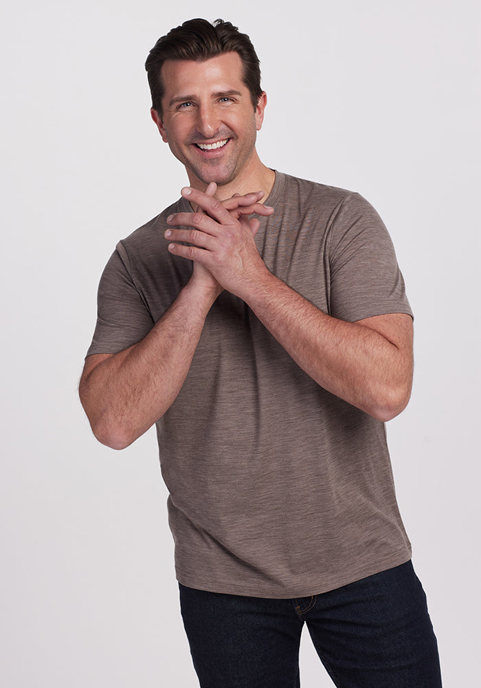 A man with short dark hair, wearing a Simply Taupe Endurance Tee by Woolx and dark jeans, smiles and clasps his hands against a plain white background. 