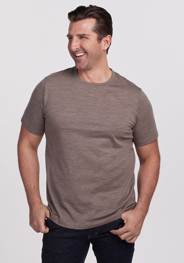 A man with short brown hair stands against a plain background, smiling. He is wearing a Simply Taupe Endurance Tee from Woolx, crafted from Merino Wool, paired with dark jeans. His hands rest casually in his pockets, exuding a relaxed vibe while highlighting his performance gear.